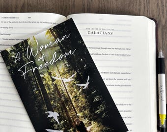 May include: A black and white book with the title "A Woman of Freedom" on the cover. The cover features a photo of a woman walking through a forest with birds flying overhead. The text "Girlfriends in the Word" and "by Natalia Drumm" are also on the cover.