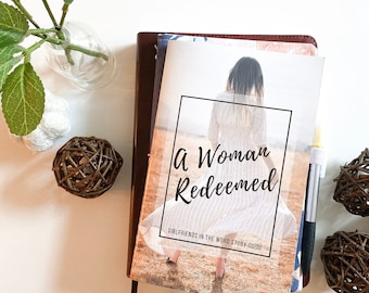 May include: A closed book with a brown cover and a white spine. The book's title is "A Woman Redeemed" with the subtitle "Girlfriends in the Word Study Guide". The cover features a woman in a white dress walking away from the camera in a field.