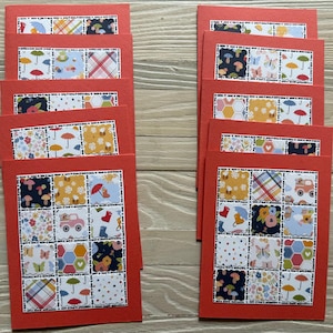 May include: A stack of red greeting cards with a patchwork design. Each card features a grid of colorful squares with patterns of umbrellas, flowers, and butterflies. The cards are arranged on a light wood surface.