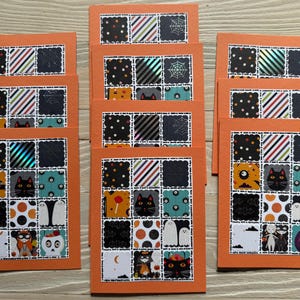 May include: A stack of orange Halloween greeting cards. Each card features a patchwork design with black cats, ghosts, and other spooky images. The cards have a white border and are arranged on a wooden surface.