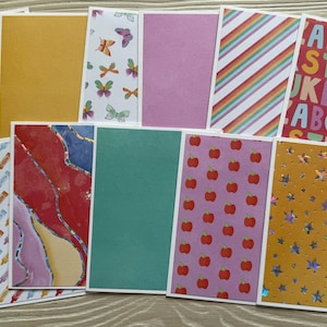 May include: A collection of colorful blank greeting cards with various patterns. Designs include butterflies, apples, crayons, rainbows, and an alphabet pattern. Some cards have text that reads "Share the year WRITE".