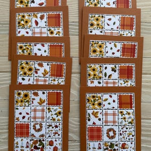 May include: A collection of fall-themed greeting cards. Each card features a brown border and a white interior with a patchwork design of sunflowers, leaves, and plaid patterns. The cards are arranged on a light wood surface.
