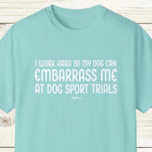 以下が含まれることがあります： 白文字で「I WORK HARD SO MY DOG CAN EMBARRASS ME AT DOG SPORT TRIALS」と書かれた、水色のTシャツ。下に「doghaus」の文字。柔らかい素材で作られたシャツです。