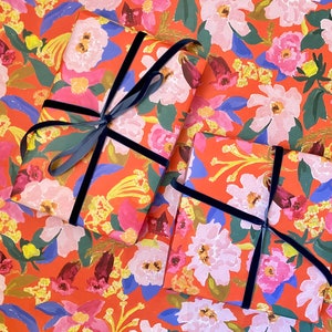 May include: Two wrapped gifts on floral wrapping paper with a vibrant orange background. The paper features a pattern of pink, white, and blue flowers with green leaves. Each gift is tied with a dark blue velvet ribbon.