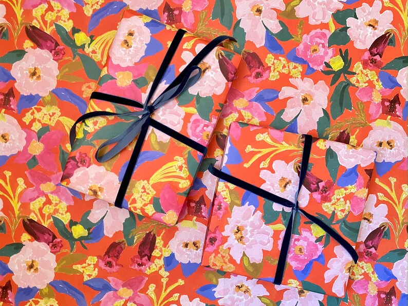 May include: Two wrapped gifts on floral wrapping paper with a vibrant orange background. The paper features a pattern of pink, white, and blue flowers with green leaves. Each gift is tied with a dark blue velvet ribbon.