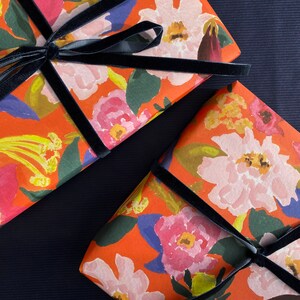 May include: Two gift boxes wrapped in orange floral paper with a black velvet ribbon. The paper features a watercolor design of pink, white, and red flowers with green leaves. The boxes are arranged diagonally on a dark blue surface.