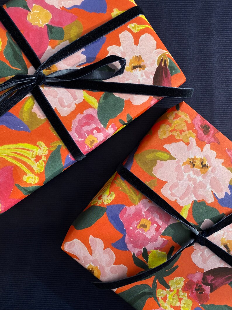 May include: Two gift boxes wrapped in orange floral paper with a black velvet ribbon. The paper features a watercolor design of pink, white, and red flowers with green leaves. The boxes are arranged diagonally on a dark blue surface.