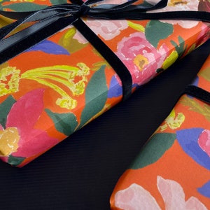 May include: Two gift boxes wrapped in floral patterned paper with a vibrant orange background. The paper features pink, yellow, and blue flowers. A black velvet ribbon is tied around each box.