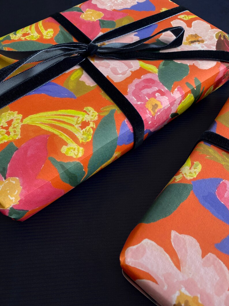 May include: Two gift boxes wrapped in floral patterned paper with a vibrant orange background. The paper features pink, yellow, and blue flowers. A black velvet ribbon is tied around each box.