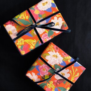 May include: Two wrapped gifts with floral patterned paper in shades of red, pink, white, and yellow. The gifts are accented with navy blue velvet ribbon and bows. The background is black.