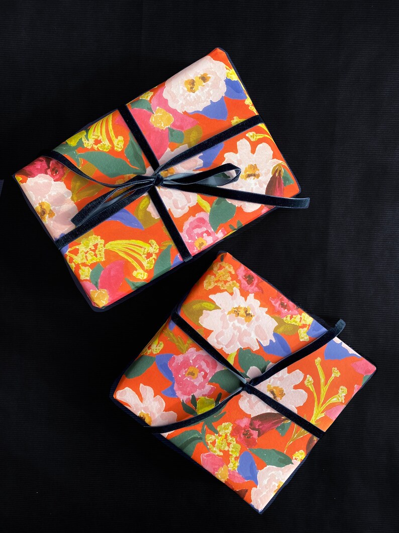 May include: Two wrapped gifts with floral patterned paper in shades of red, pink, white, and yellow. The gifts are accented with navy blue velvet ribbon and bows. The background is black.