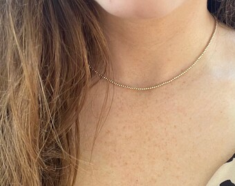 May include: A delicate gold chain necklace with a beaded design. The necklace is worn by a person with long brown hair and a light brown top with a black and white animal print.