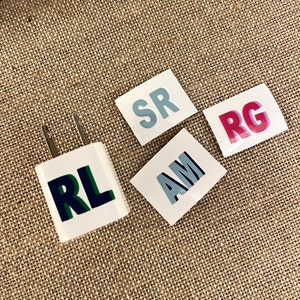 May include: Four white stickers with different colored letters on them. The stickers say "RL", "AM", "SR", and "RG". The stickers are on a brown burlap background.