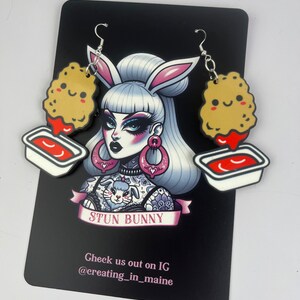 May include: A pair of dangle earrings featuring a cartoon chicken nugget with a smiling face and a small red container of dipping sauce. The earrings are hanging from a silver hook and chain. The background is a black card with the text "Stun Bunny" and "Check us out on IG @creating_in_maine".