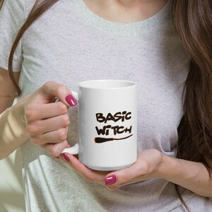 May include: White ceramic mug with the words "BASIC WITCH" in black script. The mug has a black witch's broom graphic below the text. The mug is held by a person wearing a gray t-shirt.