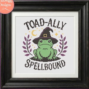 May include: A cross-stitch pattern in a black frame featuring a green frog wearing a purple witch hat. The text reads TOAD-ALLY SPELLBOUND. The design includes a yellow crescent moon, small stars, and purple leafy branches on a white background.