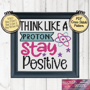 Puede incluir: Un patrón de punto de cruz con un diseño colorido con el texto "Think Like A Proton Stay Positive". El patrón está enmarcado en un marco negro con un fondo blanco.