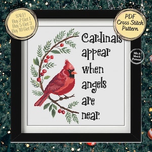 Può includere: Un modello a punto croce incorniciato con un cardinale rosso appollaiato su un ramo, bacche e il testo "Cardinals appear when angels are near". Il disegno è circondato da una ghirlanda verde.