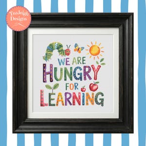 Peut inclure: Une œuvre d'art au point de croix encadrée avec le texte "WE ARE HUNGRY FOR LEARNING". Le motif présente une chenille colorée, un papillon, un soleil et une pomme. Le cadre est noir et le fond est blanc.