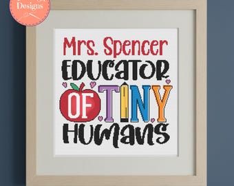Op de afbeelding: Een ingelijst kruissteekontwerp met de tekst "Mrs. Spencer EDUCATOR OF TINY HUMANS." Het ontwerp bevat een rode appel, potloden en harten. De lijst is lichtbruin en de achtergrond is wit.
