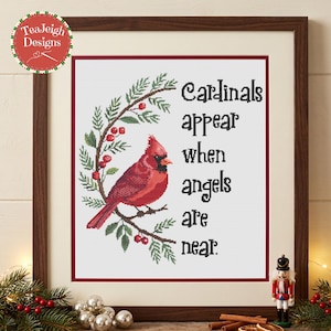 May include: A framed cross-stitch artwork featuring a red cardinal perched on a pine branch with red berries. The text reads, Cardinals appear when angels are near. The design is set against a white background with a dark red border.
