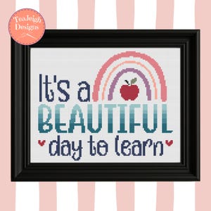 May include: A framed cross-stitch piece with the text "It's a BEAUTIFUL day to learn" and a rainbow design. The rainbow is in pastel colors, and a red apple is in the center. The frame is black.