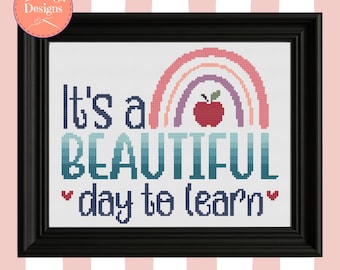 Op de afbeelding: Een ingelijst kruissteekwerk met de tekst "It's a BEAUTIFUL day to learn" en een regenboogontwerp. De regenboog is in pastelkleuren en een rode appel staat in het midden. De lijst is zwart.