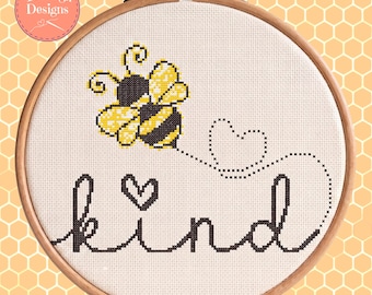 Può includere: Ricamo a punto croce in un telaio di legno con un'ape gialla e nera e una scia di cuori tratteggiati. La parola "kind" è ricamata sotto, con un cuore sopra la "i". Il disegno è su tessuto bianco.