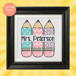 May include: Framed cross-stitch art featuring three colorful pencils with polka dot and chevron patterns. The pencils are teal, pink, and purple, with the name "Mrs. Peterson" in the center. The artwork is framed in a black frame.