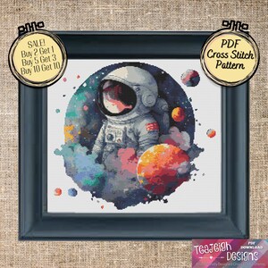 May include: A cross stitch pattern featuring an astronaut in a white spacesuit standing in front of a colorful nebula with planets. The pattern is framed in a black frame and has a white background. The text "PDF Cross Stitch Pattern" is on a yellow circle in the upper right corner of the image.
