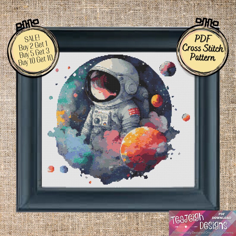 May include: A cross stitch pattern featuring an astronaut in a white spacesuit standing in front of a colorful nebula with planets. The pattern is framed in a black frame and has a white background. The text "PDF Cross Stitch Pattern" is on a yellow circle in the upper right corner of the image.