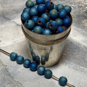 May include: A collection of blue, glittery beads in a small silver bucket, with a strand of beads strung on a wire. The beads have a textured surface and a metallic sheen, with hints of green and purple. The beads are round and appear to be about 0.5 inches in diameter.
