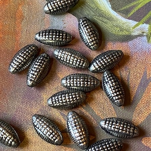 May include: A collection of silver-colored, oblong beads with a textured, grid-like pattern. The beads are scattered on a background with muted tones of brown, green, and purple.