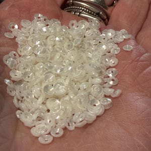 May include: A handful of translucent, iridescent, flower-shaped beads. The beads are small, with a hole in the center, and appear to be made of plastic or a similar material. A silver crown ring is visible in the background.