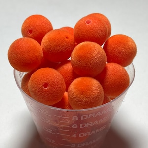 May include: A clear plastic measuring cup filled with orange fuzzy balls. The balls have a small hole in the center. The cup has measurements in fluid ounces and drams.