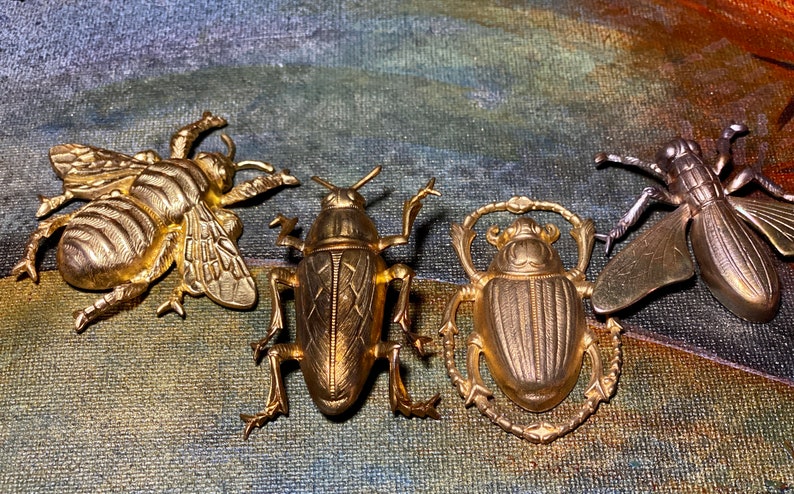 May include: Four detailed, gold-toned insect brooches. The collection includes a bee, a beetle, a scarab beetle, and a fly. Each brooch is intricately designed with realistic features, showcasing the insects' wings, legs, and body segments. The brooches are displayed against a textured background.