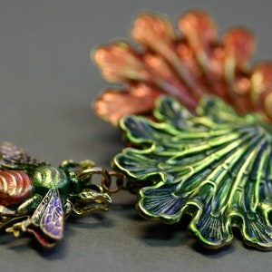 May include: A detailed, colorful bee pendant with a matching green and orange leaf-like charm. The bee features red, green, and purple accents. The jewelry is made of metal and has a vintage aesthetic.