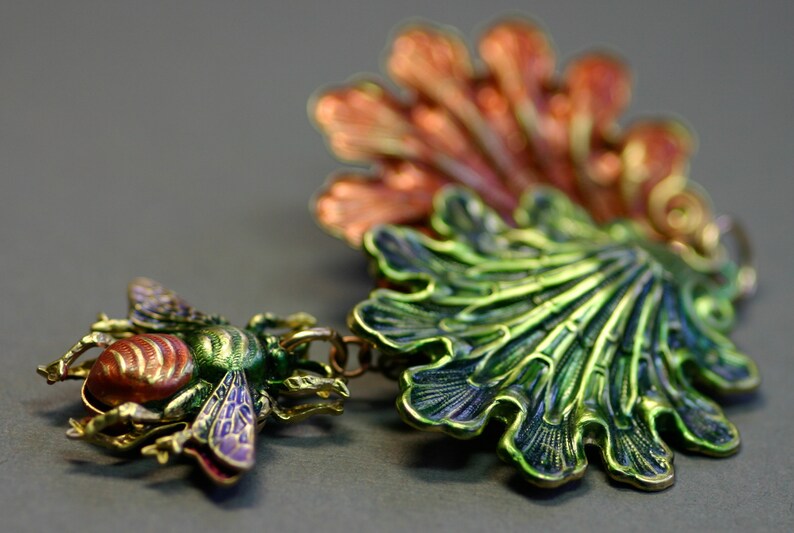 May include: A detailed, colorful bee pendant with a matching green and orange leaf-like charm. The bee features red, green, and purple accents. The jewelry is made of metal and has a vintage aesthetic.