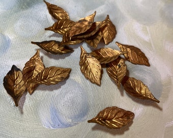 May include: A collection of small, gold-colored metal leaf embellishments. Each leaf has detailed veining and a slightly curved shape. The leaves are scattered on a light blue and white painted background, creating a natural, decorative theme.