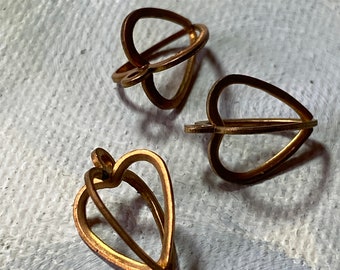 May include: Three gold-colored heart-shaped pendants. Each pendant features a delicate, openwork design, with the heart shape formed by thin, curved metal lines. The pendants are arranged on a textured, light-colored surface.
