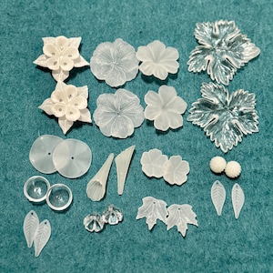May include: A collection of translucent white floral and leaf-shaped embellishments. The assortment includes various flower designs, leaves, and other decorative elements, perfect for crafting projects. The pieces are arranged on a teal surface.