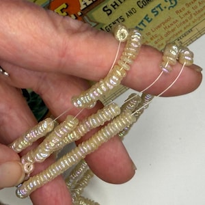 May include: Multiple strands of small, iridescent, cream-coloured disc-shaped beads strung on thin white thread. The beads have a glossy, pearlescent finish that reflects light, creating a subtle rainbow sheen across their circular, flat surfaces.