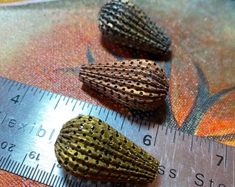 May include: Close-up of several metal beads with a textured, perforated design. The beads are teardrop-shaped and come in various metallic colors, including gold, bronze, and copper. A metal ruler is in the foreground, providing scale.