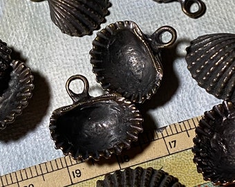 May include: Close-up of several small, bronze-colored seashell charms with attached loops. The charms have a detailed, ridged texture and are arranged on a light-colored surface. The shells are open and closed, showing their interior.