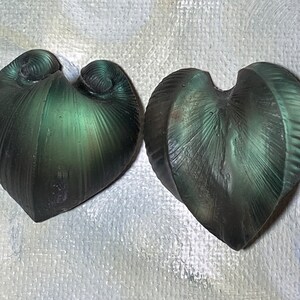 May include: Two heart-shaped, dark green objects with a textured, ridged surface. The objects have a swirling design at the top and a pointed bottom. The color shifts slightly, creating a subtle iridescent effect. The objects appear to be decorative.