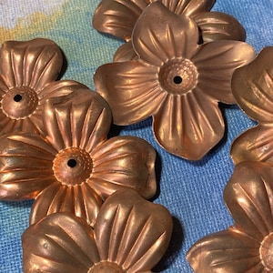 Può includere: Primo piano di diversi abbellimenti floreali in metallo color rame. Ogni fiore ha cinque petali e un piccolo foro al centro. I fiori sono disposti su una superficie blu e bianca.