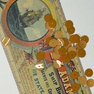 May include: A collection of small, round, mustard-yellow sequins scattered across a vintage-style paper advertisement featuring a sailing ship illustration and a ruler with markings in inches, measuring up to 24 inches in length.