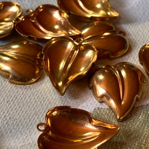 May include: Close-up of several gold-colored leaf-shaped charms. Each charm has a detailed vein pattern and a small loop at the top for attaching to jewelry or other crafts. The charms are scattered on a textured surface.