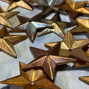 May include: A collection of metallic star-shaped embellishments in various shades of gold and silver. Each star has a textured surface and a small hole in the center, suggesting they are designed for sewing or crafting projects. The stars are scattered across a light-colored surface.