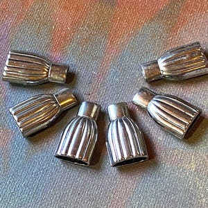 May include: Six silver-colored metal bead end caps. The caps have a ribbed, rounded design on the main body and a cylindrical opening. The caps are arranged in a circular pattern on a multicolored surface.